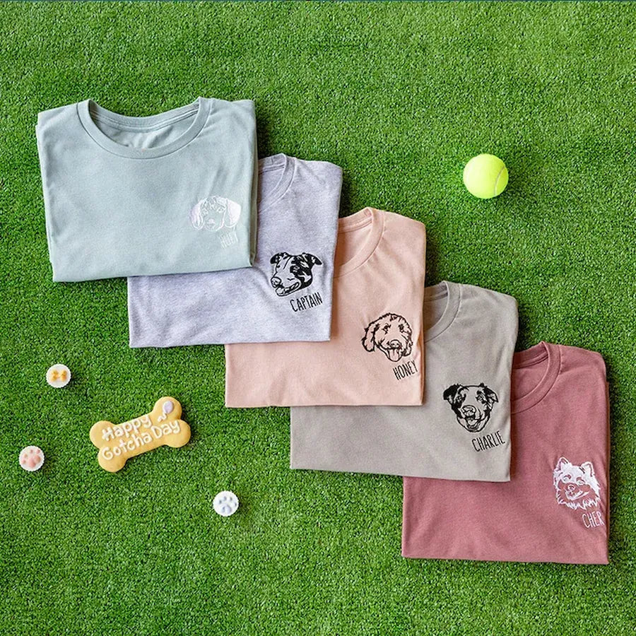 Five pastel pet portrait T-shirts arranged on grass with dog toys