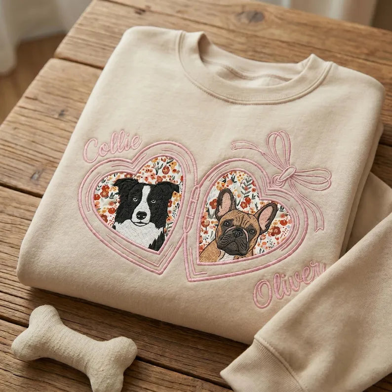 Cream sweatshirt with two dog portraits inside pink floral heart lockets on a wood table
