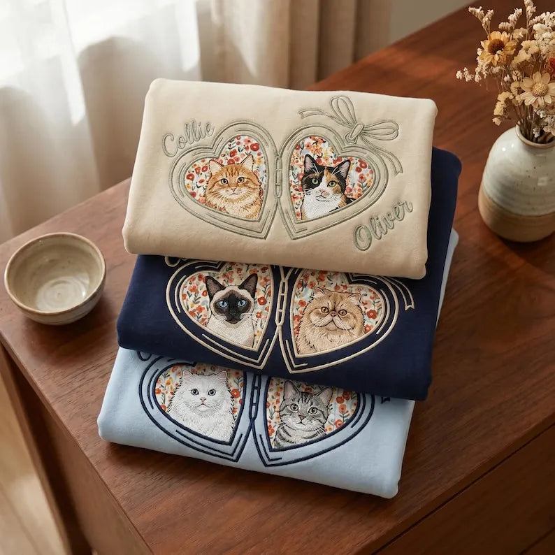 Three folded pet portrait heart locket sweatshirts beside a cup and flowers