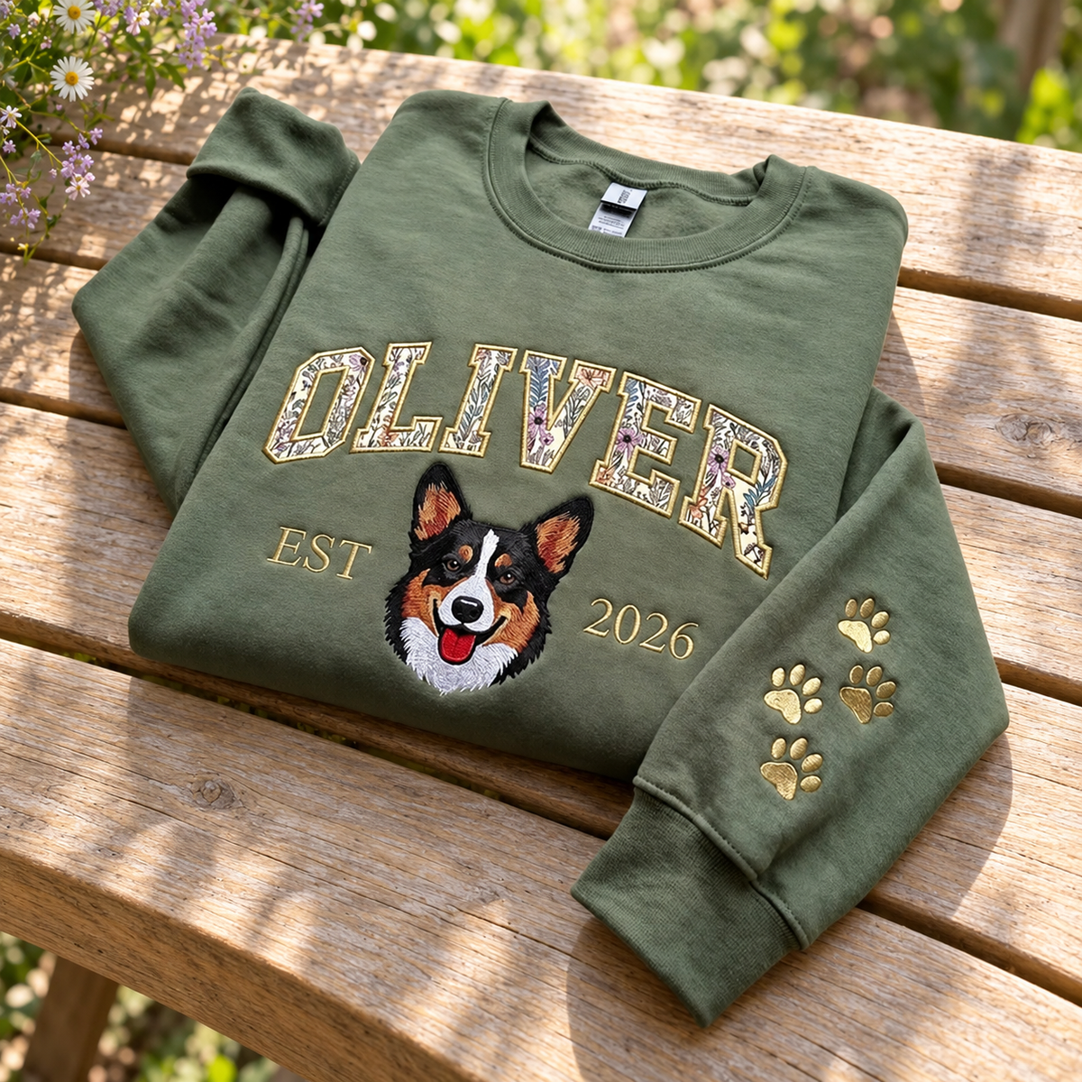 Olive OLIVER floral applique corgi sweatshirt with EST 2026 and gold paw prints on a sunlit garden bench