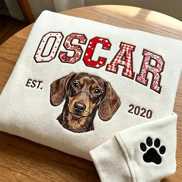 Cream sweatshirt with OSCAR in red patchwork fabric letters above an embroidered dachshund and EST 2020, paw on the cuff