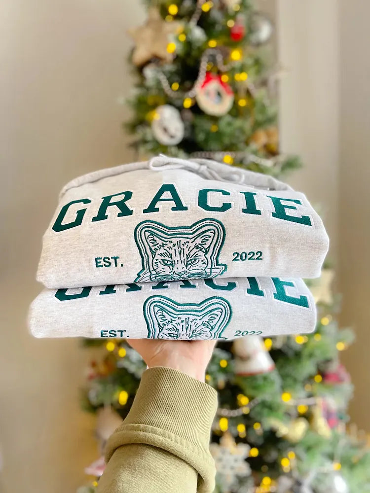 Two folded gray sweatshirts with GRACIE in green varsity letters and a cat outline