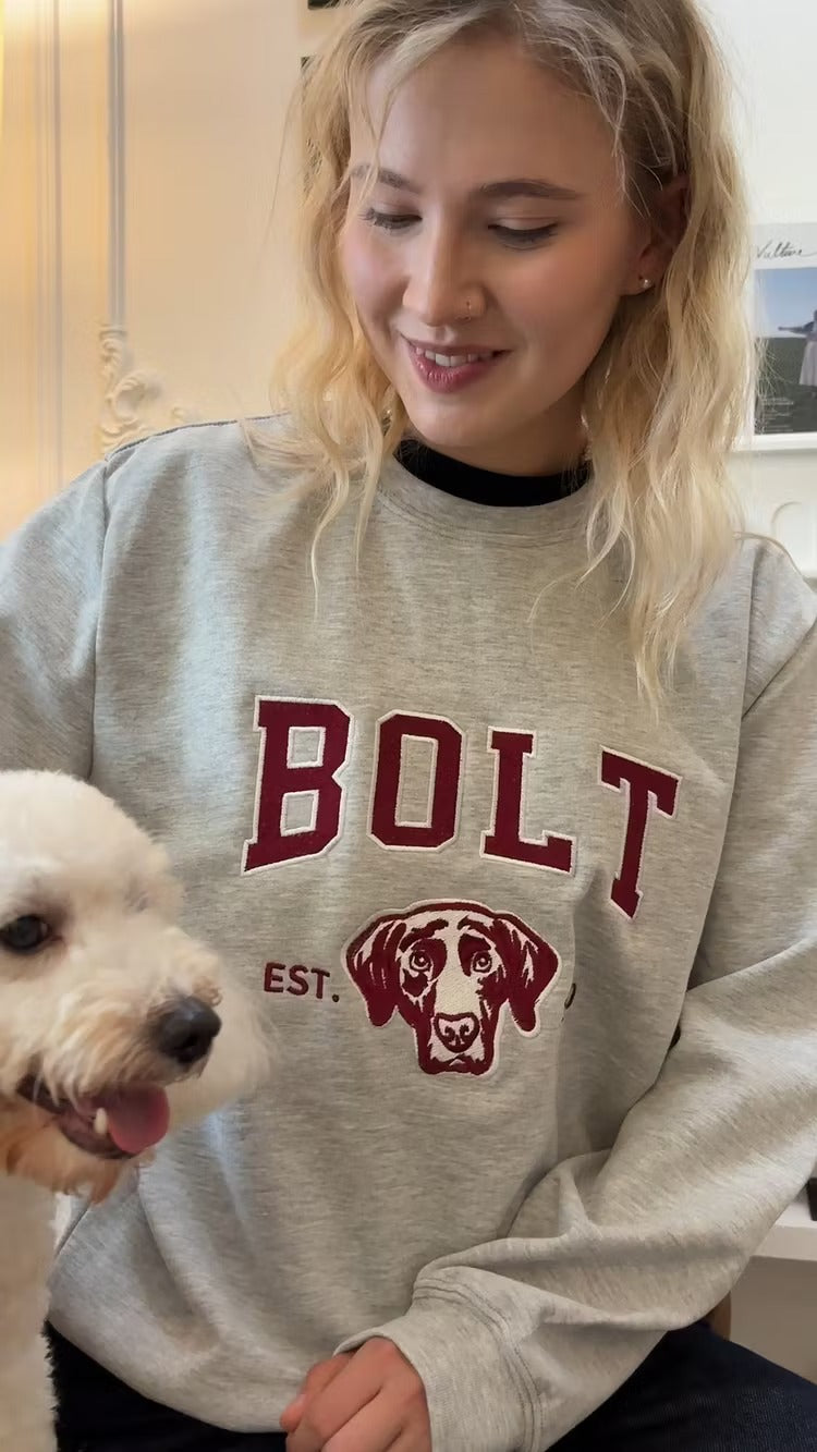 Woman in a gray sweatshirt with BOLT in maroon letters and a Labrador outline, next to a white poodle