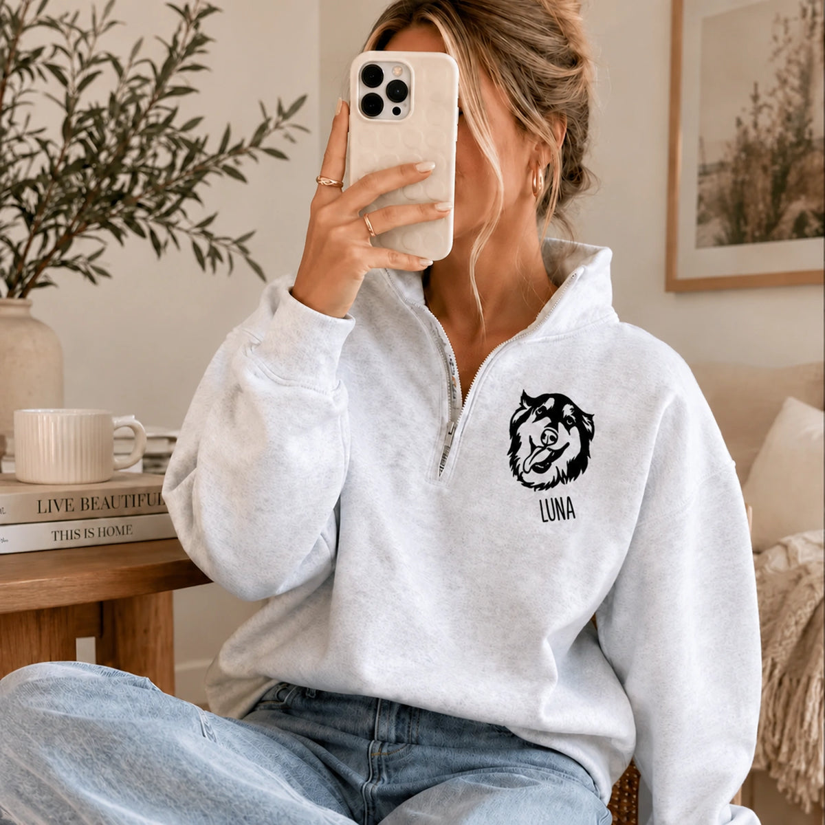 Woman taking a mirror selfie in a white quarter zip sweatshirt with a line art dog portrait and the name LUNA